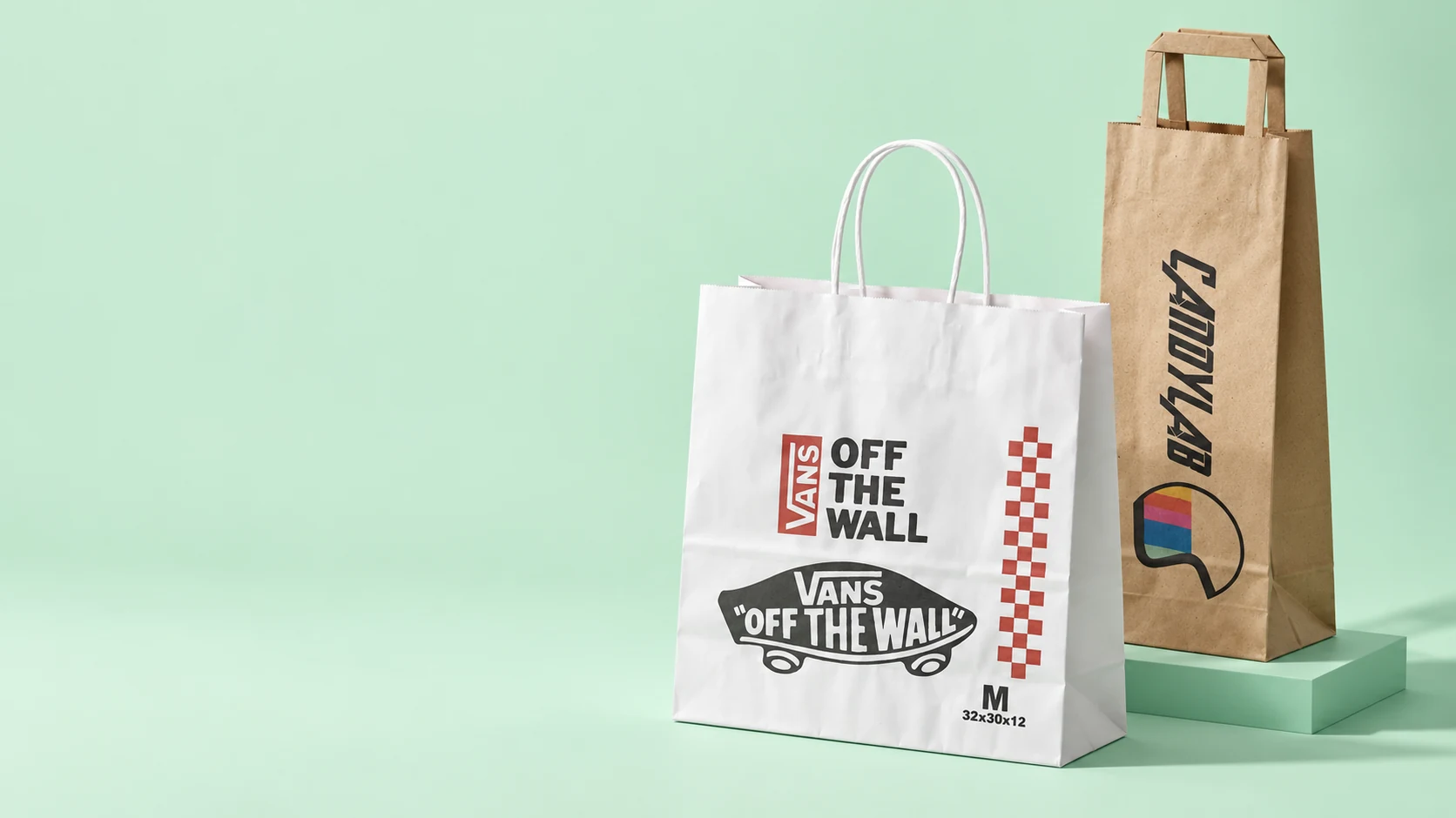 Bolsas de papel personalizadas: blanca con asas torcidas y kraft con asas planas, en una escena editorial sobre fondo menta